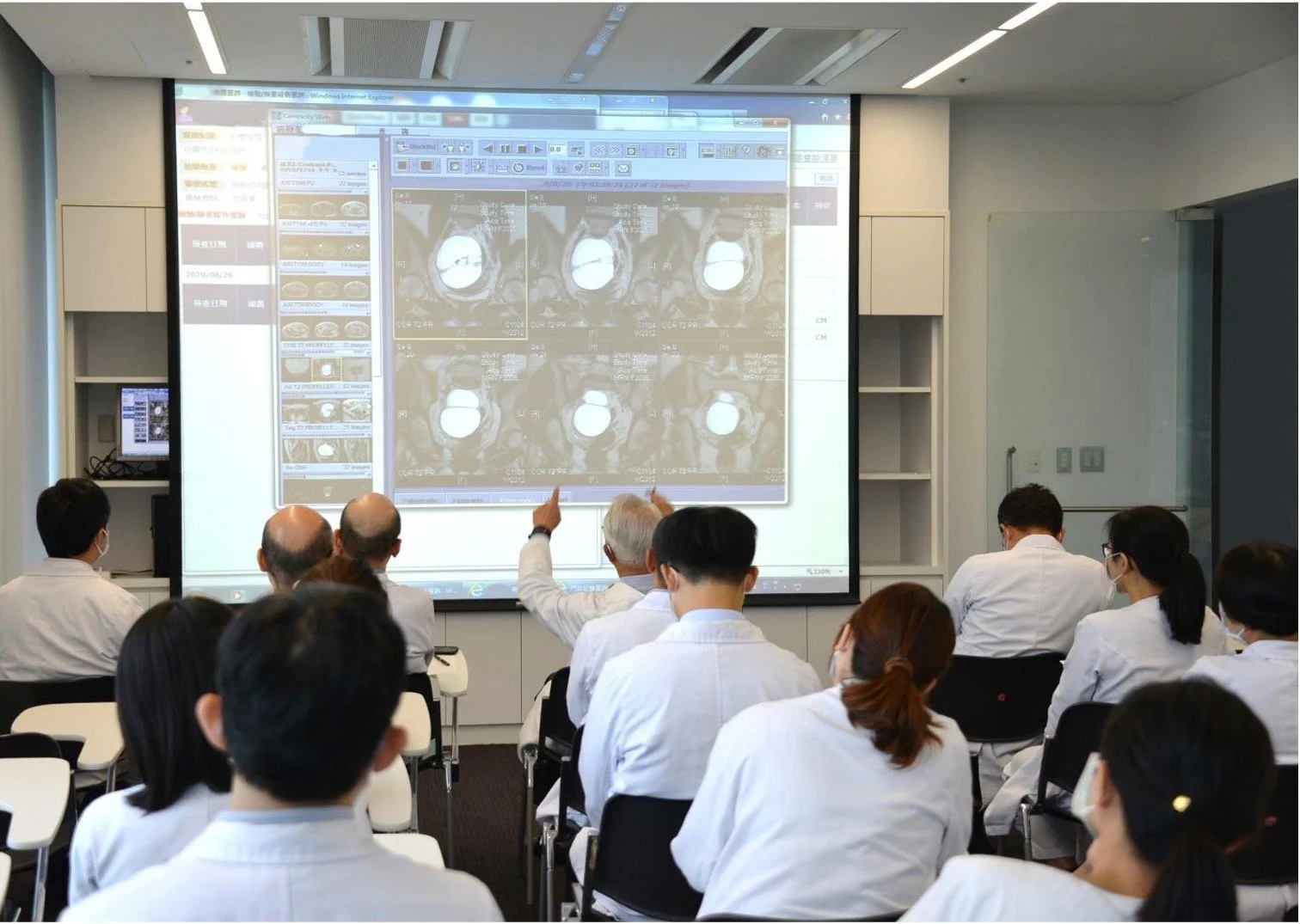A group of medical professionals, dressed in white lab coats, attending a presentation of medical scans projected onto a large screen in a conference room.