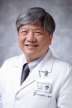 A smiling man in a white lab coat with a badge, standing against a gray background.