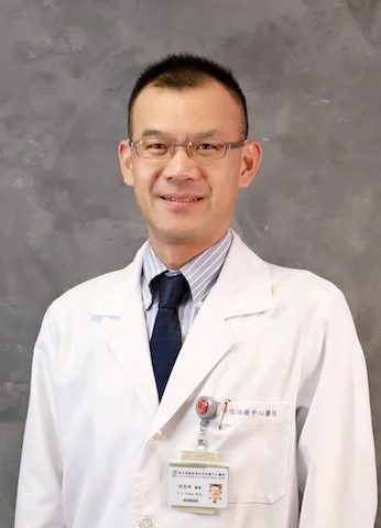 A male doctor wearing glasses, a white lab coat, a light blue shirt, and a dark tie, smiling at the camera against a gray background.