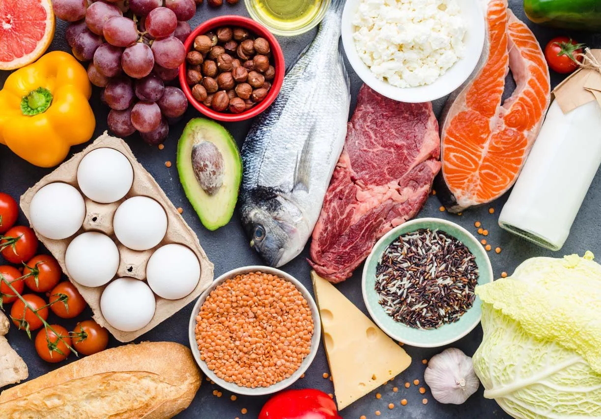 Assorted fresh foods including grapes, grapefruit, yellow bell pepper, eggs, avocado, fish, ground beef, salmon fillet, milk, cherry tomatoes, bread, lentils, rice, cheese, garlic, lettuce, and various condiments on a dark surface.