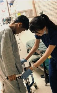 A young man with a walker in a hospital gown and a nurse assisting him, both wearing gloves, in a hospital room.