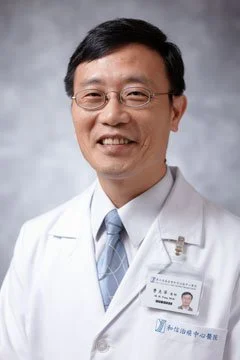 A male doctor wearing glasses, a white lab coat with a name badge, and a light blue tie, smiling in front of a gray background.