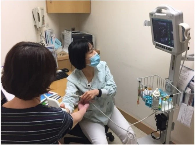 A woman with brown hair and a striped shirt assists a woman with black hair wearing a blue face mask, sitting in a medical exam room, during an ultrasound scan. The patient is holding an ultrasound probe while a monitor displays the scan. Medical equ