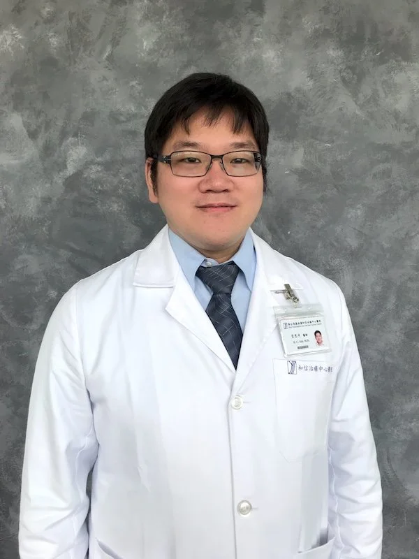 A male doctor in white coat, wearing glasses, a light blue shirt, and a tie, standing in front of a gray textured background.