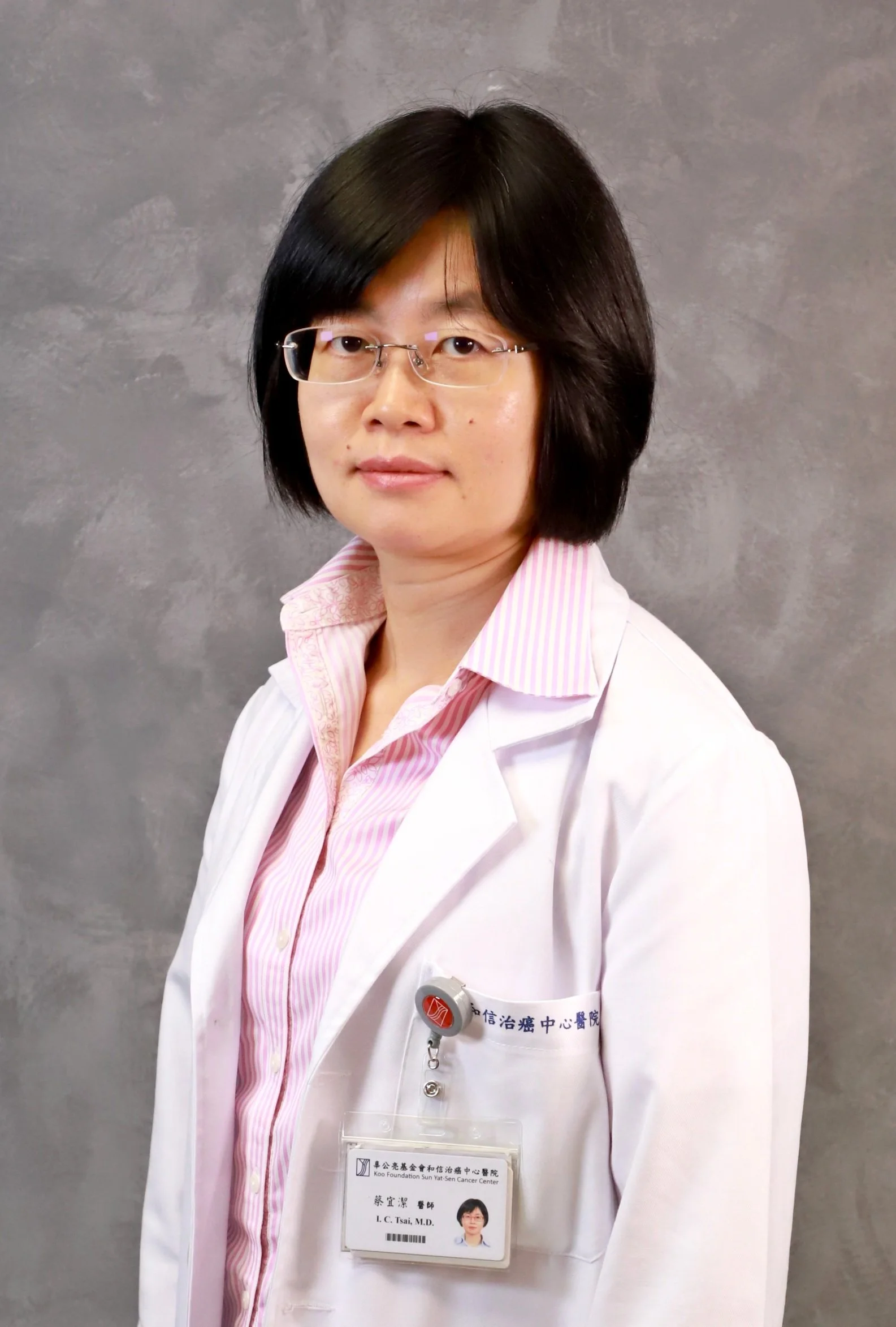 A woman with glasses and short black hair, wearing a white lab coat and pink striped shirt, posing against a gray background.