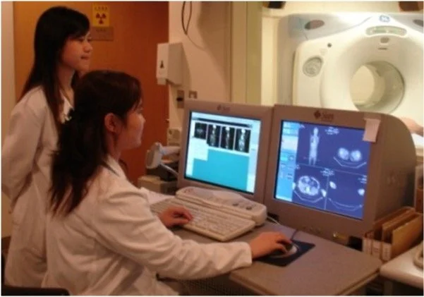 Two medical professionals, a woman and a girl, in a laboratory working on a computer displaying medical imaging scans, with MRI or CT scanner visible in the background.