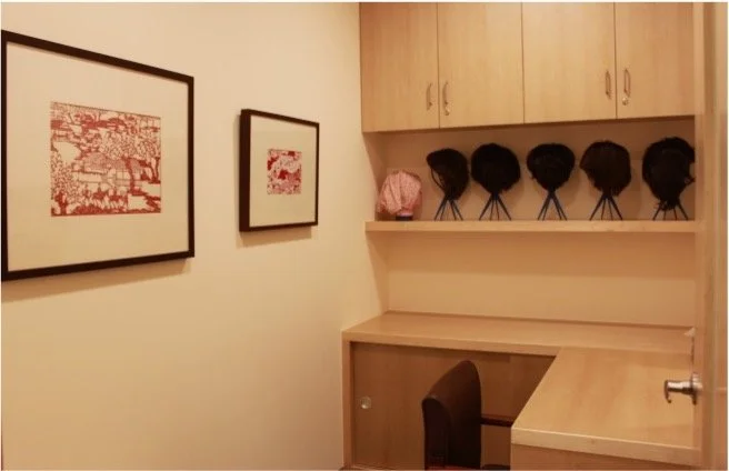 A corner of a room with framed art on the wall, a desk with a chair, and six hats stored on a shelf above the desk.