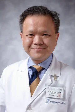 A male doctor in a white coat, wearing a name badge, smiling at the camera against a gray background.