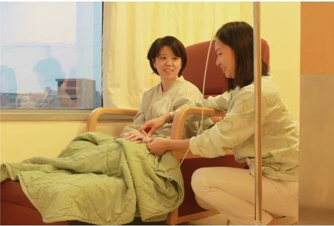 A woman in a hospital bed connected to IV therapy, conversing with a healthcare worker or visitor sitting beside her, both smiling.