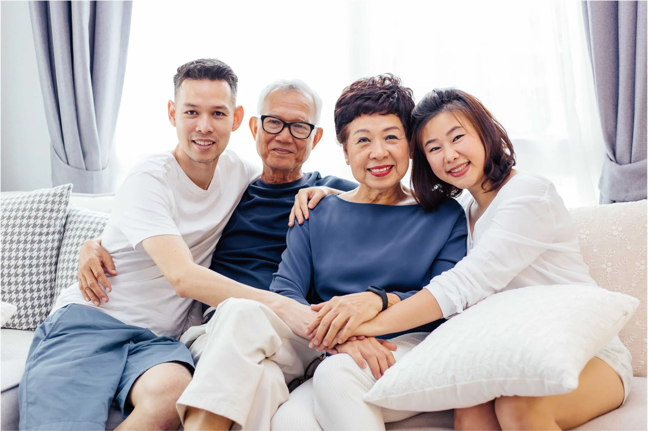A multigenerational Asian family sitting on a sofa together, smiling, with a bright window and curtains in the background.