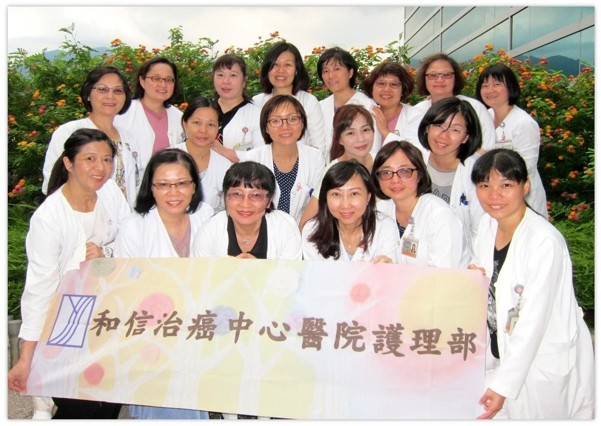 Group of healthcare professionals, mostly women, wearing white coats, gathered outdoors in front of flowering bushes, holding a banner with Chinese characters.