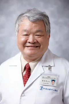 Portrait of an older man in a white doctor's coat, smiling, with gray hair and a name badge, against a gray background.