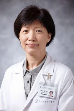 A woman in a white medical coat with a name badge, standing against a grey background.
