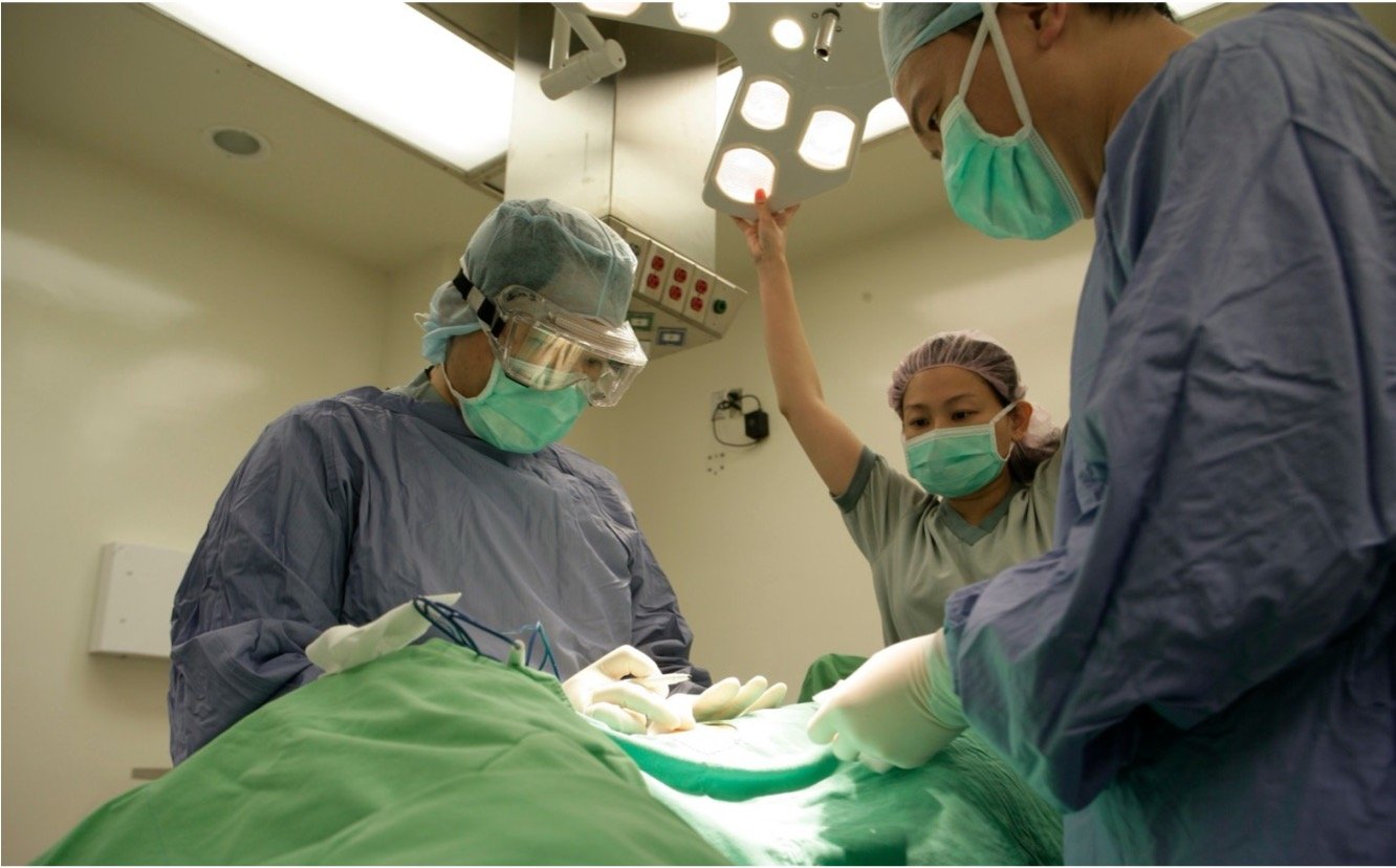 Medical team performing surgery in an operating room, with surgeons wearing surgical scrubs, masks, and gloves, and a nurse or assistant monitoring overhead lights.