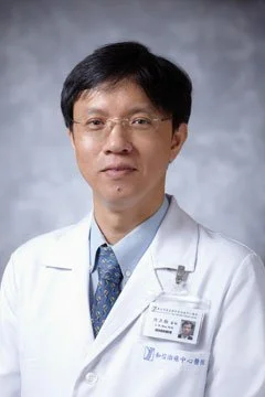 A male doctor wearing glasses and a white lab coat, standing against a gray background.