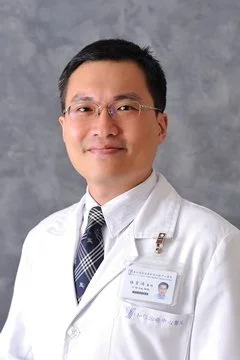 A male doctor in a white coat, wearing glasses, with a name tag, smiling against a gray background.