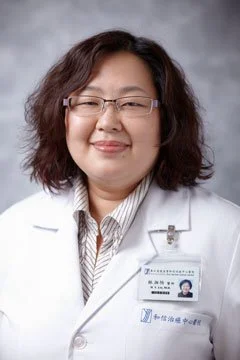 Portrait of a woman wearing glasses and a white medical coat with a name tag, smiling against a gray background.