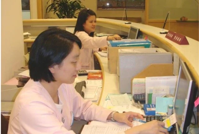 Two women working at a curved office reception desk with files, papers, and office supplies.