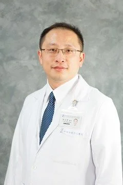 A man in a white medical coat and glasses standing against a gray background.