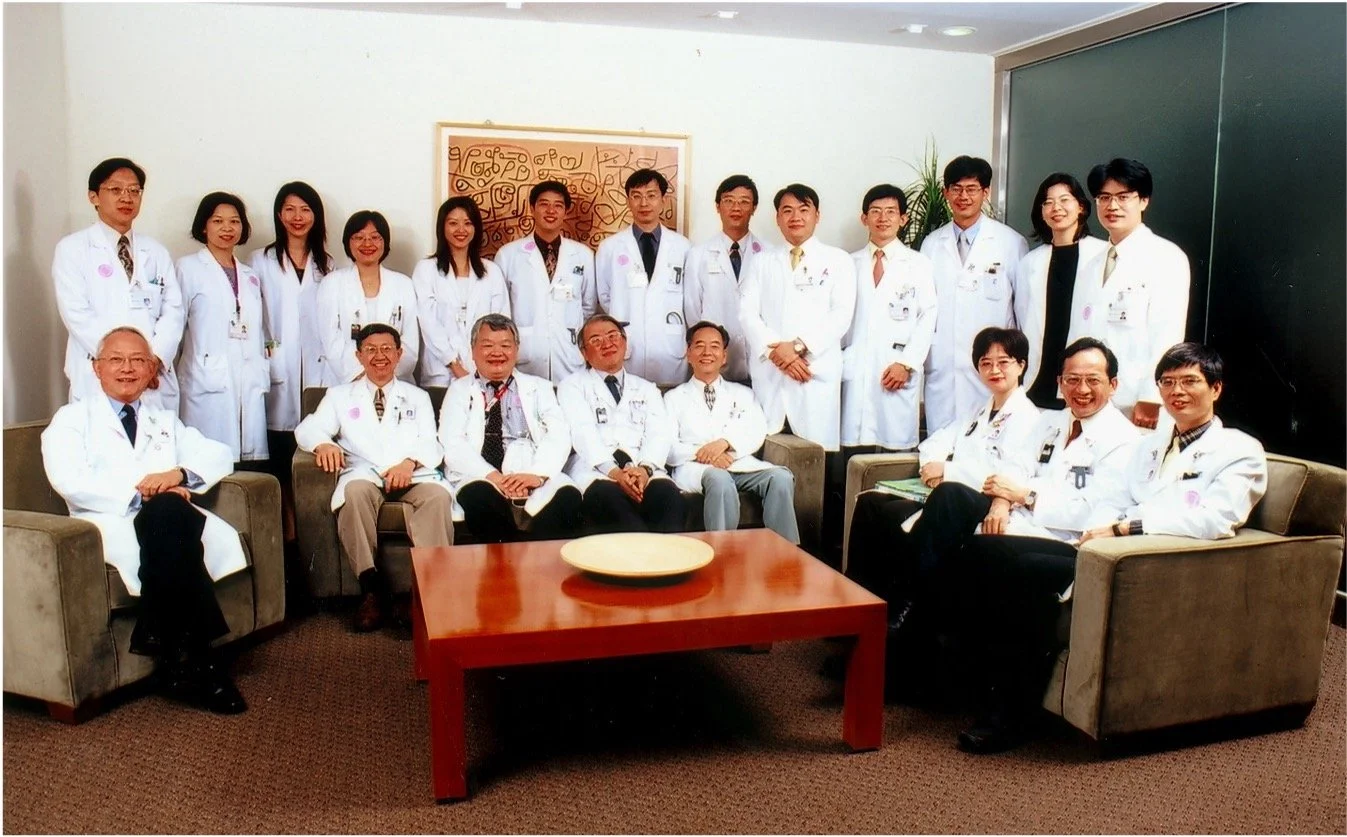 A group of medical professionals, mostly doctors, gathered in a conference room. They are dressed in white coats and are seated and standing around a table and couches, smiling for the photo.