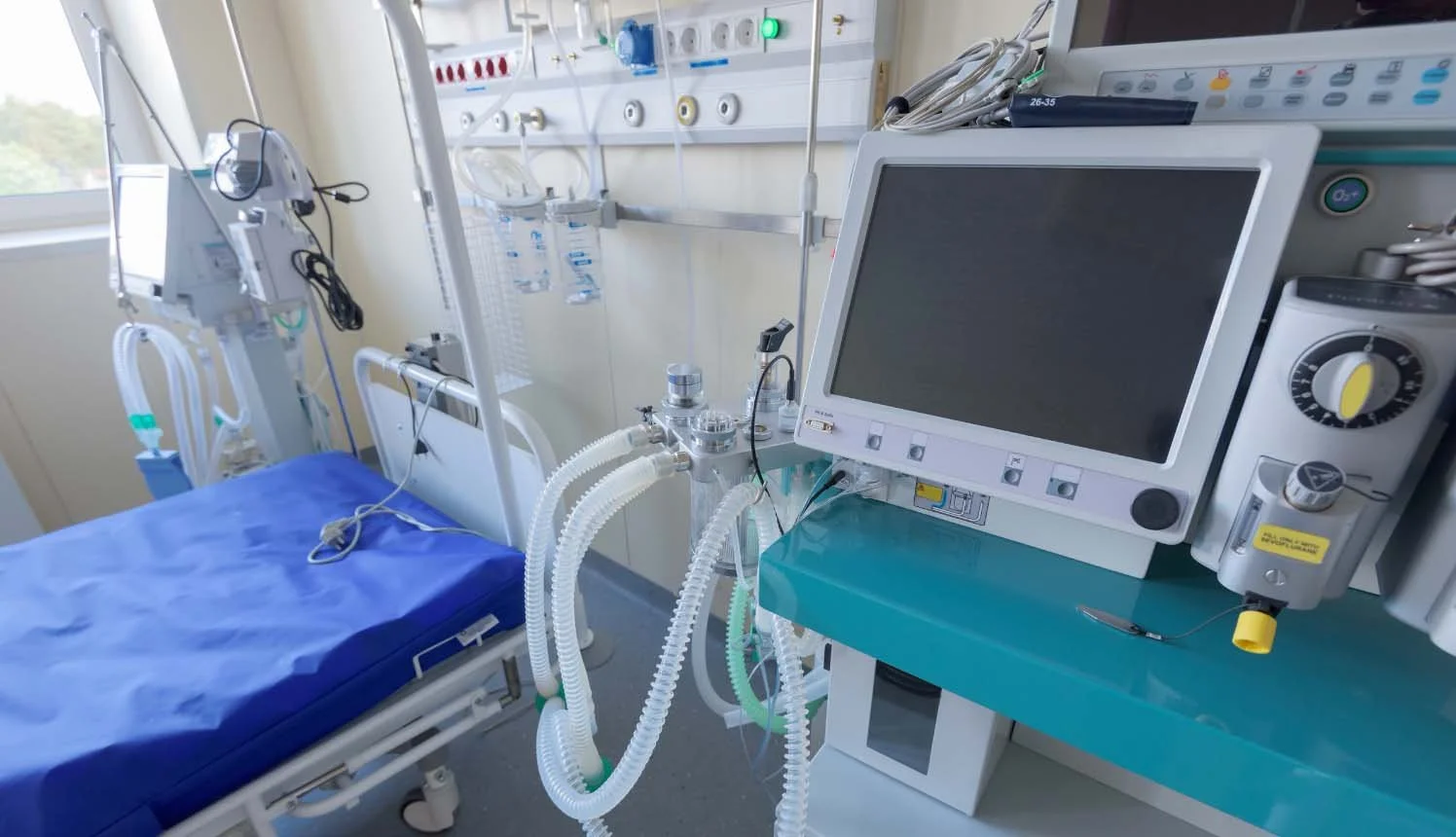 Hospital ICU room with medical equipment, a hospital bed with blue sheets, monitors, ventilator, and IV supplies.