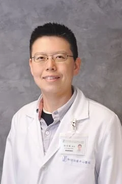 A man wearing glasses and a white lab coat, smiling against a plain gray background.