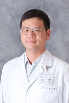 Portrait of a man in a white medical coat with glasses, standing in front of a gray background.