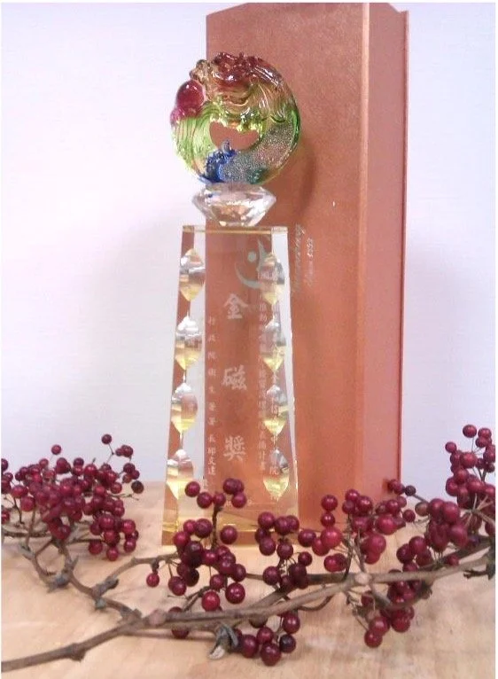 A decorative glass ball with colorful swirls sits atop a tall, clear, faceted crystal award or trophy, with a pink box in the background and red berries with a branch in the foreground.
