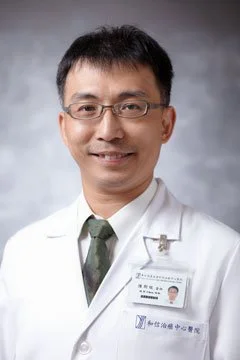 A male doctor wearing glasses, a white lab coat, and a name badge, smiling against a neutral background.
