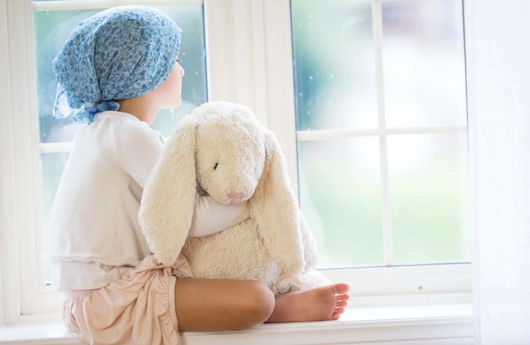 A young child with a blue headscarf sitting cross-legged on a windowsill, holding a large plush bunny, looking out the window.