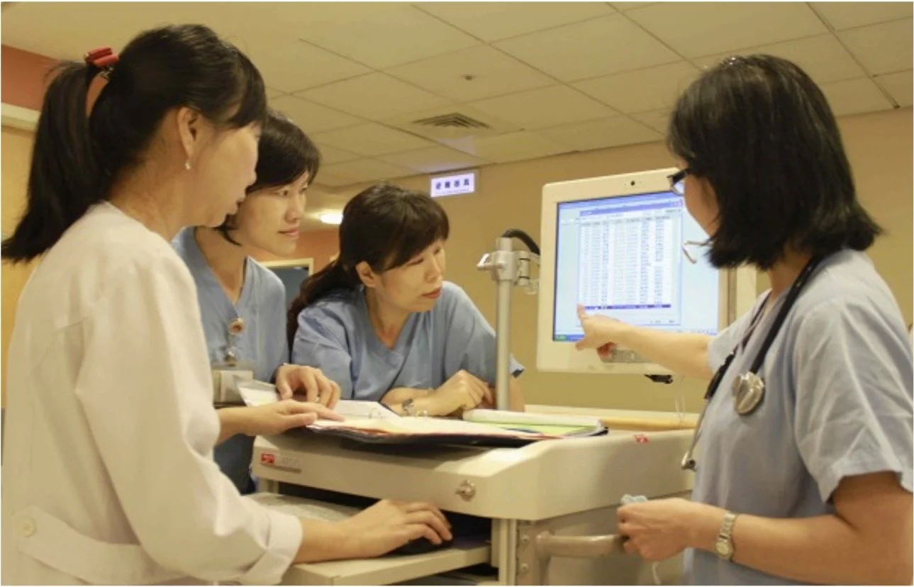 Four nurses or healthcare professionals gathered around a monitor in a hospital or medical setting, reviewing data or patient information on the screen.