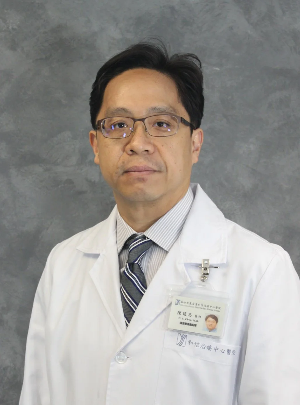 A male doctor wearing glasses, a white lab coat, and a striped tie, standing against a gray background.