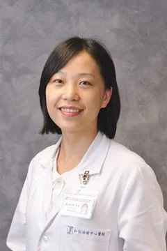 A woman in a white medical coat smiling against a gray background.