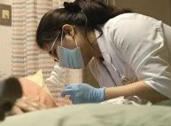 A healthcare worker wearing a mask and gloves leaning over a patient in a hospital bed.