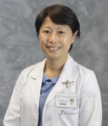 Asian female doctor in white coat smiling for a portrait. She wears a name badge and light blue shirt, standing against a gray background.