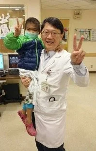 A doctor in a white coat holding a young girl, both making peace signs with their hands and wearing face masks in a hospital setting.