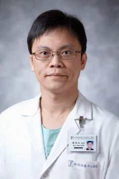 A man in a white medical coat with glasses, standing against a gray background, wearing a name badge.