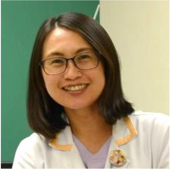 A smiling woman with shoulder-length dark hair, glasses, and a white lab coat with an orange trim, standing in front of a green and yellow wall.