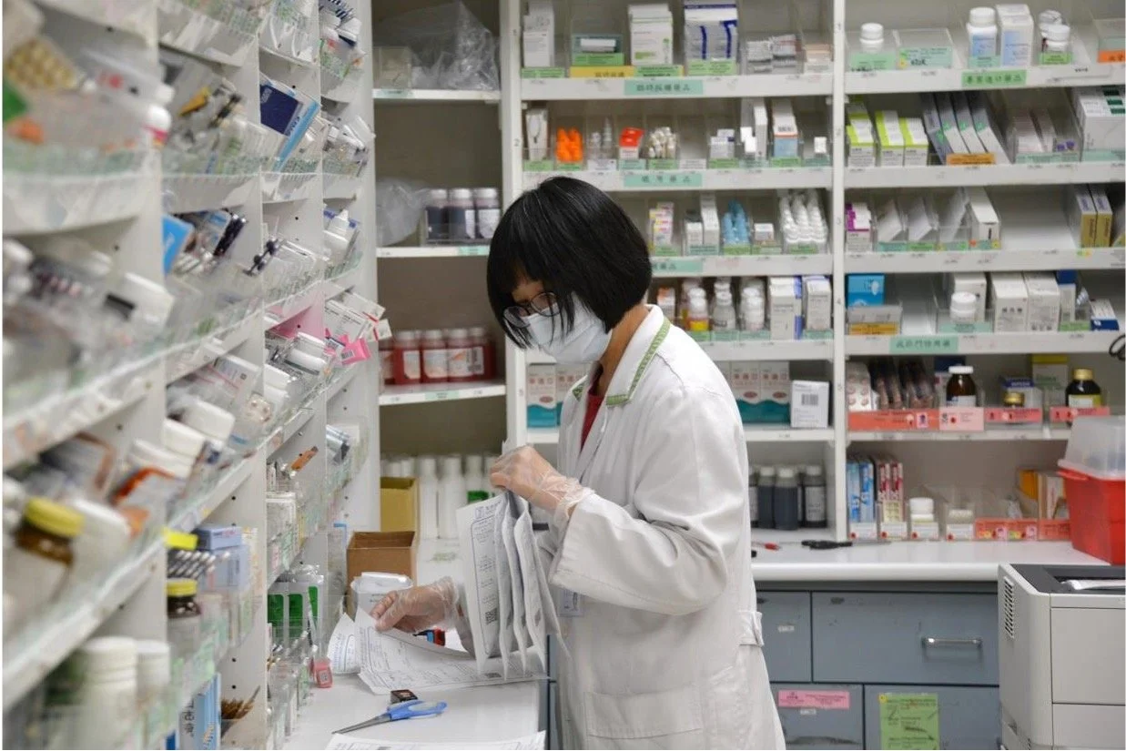 A pharmacist wearing a face mask and gloves organizing prescriptions in a pharmacy with shelves of medications in the background.