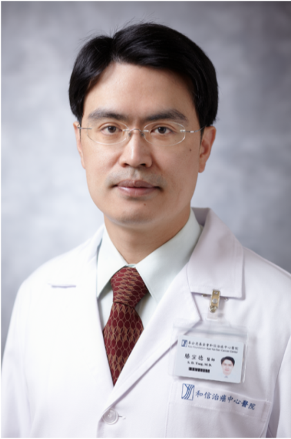 A male doctor wearing glasses and a white coat with a name badge, standing against a gray background.
