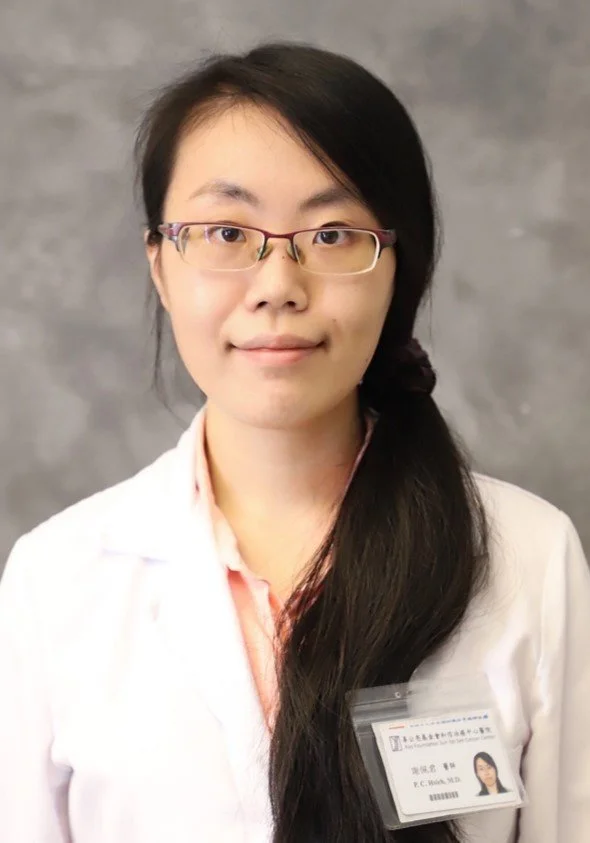 Young woman with long dark hair and glasses wearing a white coat and a name badge, standing against a gray background.