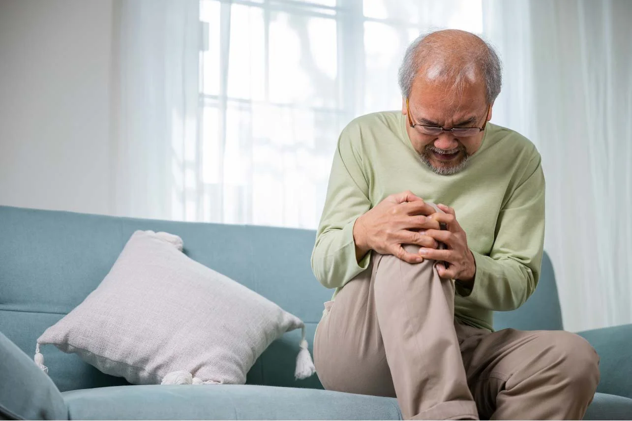 An older man sitting on a blue sofa, clutching his knee in pain, with a distressed facial expression.