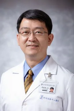 A male doctor wearing glasses, a white lab coat with a name badge, a blue shirt, and a yellow tie, smiling in a professional portrait.