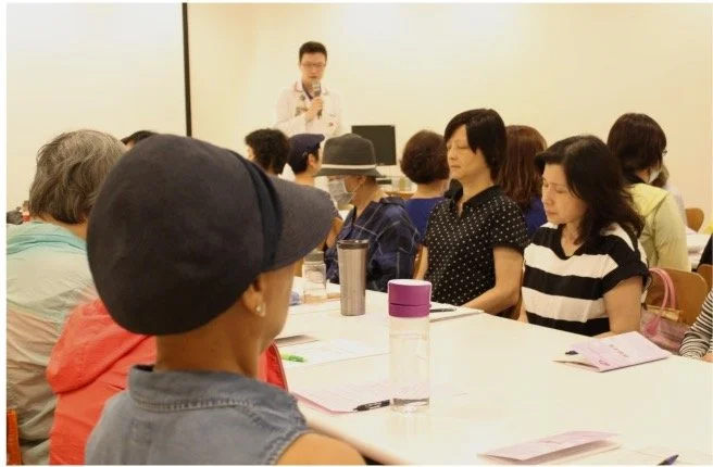 Group of people seated around a conference table listening to a speaker standing at the front of the room.