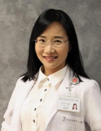 Portrait of a woman with black hair and glasses, wearing a white medical coat with a name tag, standing in front of a gray background.