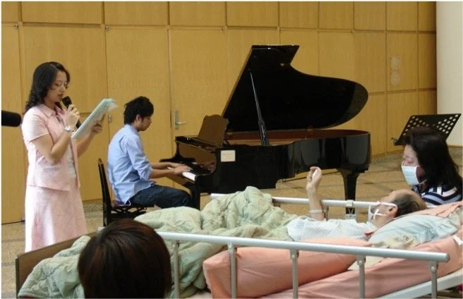 A woman reads from a paper while another woman plays the piano, with a patient lying in a hospital bed nearby, wearing a mask and glasses.