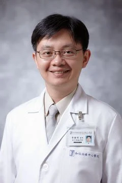 A male doctor with glasses, wearing a white coat and a name badge, smiling at the camera against a gray background.