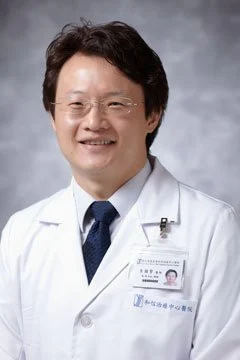 A smiling Asian male doctor wearing glasses, a white lab coat, and a blue tie posing against a gray background.