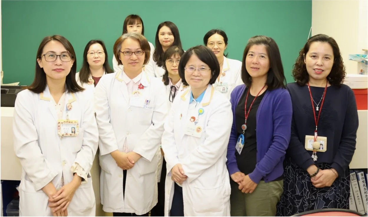 Group of female medical professionals posing in a hospital or clinic setting, some wearing white lab coats and ID badges, with a green chalkboard in the background.