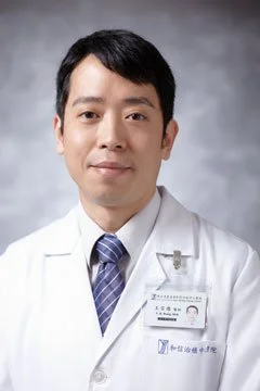 Portrait of a male doctor wearing a white lab coat and a name badge, standing against a gray background.